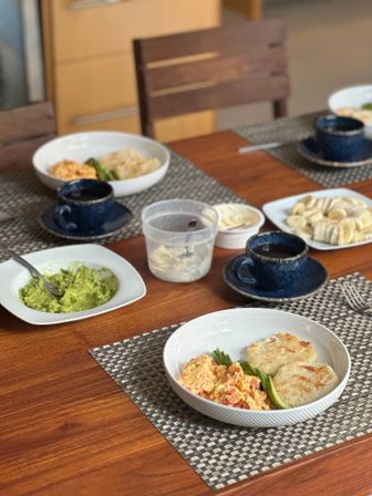 Cozy home breakfast table with bowls of scrambled eggs, grilled corn cakes and avocado, plate of sliced bananas, a dish of mashed avocado, coffee cups on a wooden table.