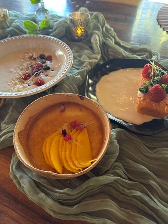 Top-down view of a dessert brunch on a wooden table with green gauze cloth: a coconut bowl of creamy mango custard topped with sliced mango and edible flowers, a white bowl of berry-topped creamy bowl, and a pastry with cream and raspberries next to a lit votive candle.