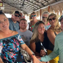 Smiling group of adults on a thatched‑roof tiki boat bar, wearing colorful summer clothes and sunglasses, holding beers for a coastal vacation vibe.