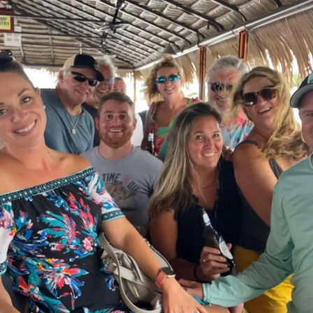 Smiling group of adults on a thatched‑roof tiki boat bar, wearing colorful summer clothes and sunglasses, holding beers for a coastal vacation vibe.
