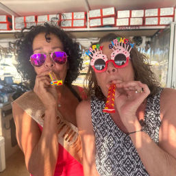 Two women wearing sunglasses and birthday glasses blowing colorful party horns at a sunny beachside bar.