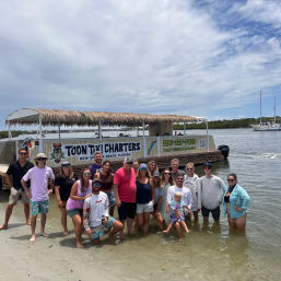 Mixed-age group posing waist-deep in shallow water beside a thatched-roof tiki party boat at New Smyrna Beach, Florida, with sandy shoreline, cloudy sky and sailboats in the background.