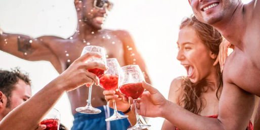 Sunlit summer beach party — a group toasting with stemmed glasses of red drink, laughing and splashing water in celebration.
