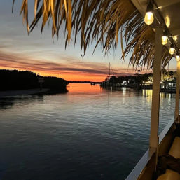 Tropical waterfront sunset over a calm inlet, orange-pink sky reflecting on water, silhouetted docks and sailboat mast, palm-frond thatched roof and warm string lights on a marina deck.