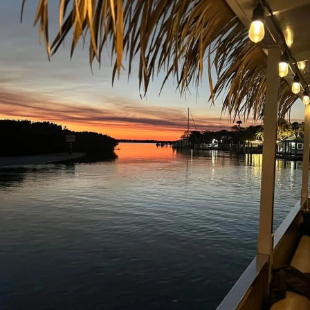 Tropical waterfront sunset over a calm inlet, orange-pink sky reflecting on water, silhouetted docks and sailboat mast, palm-frond thatched roof and warm string lights on a marina deck.