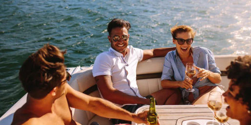 Four friends in sunglasses laughing and toasting with wine and beer on a sunlit motorboat cruising on open water, summer boat party vibe.