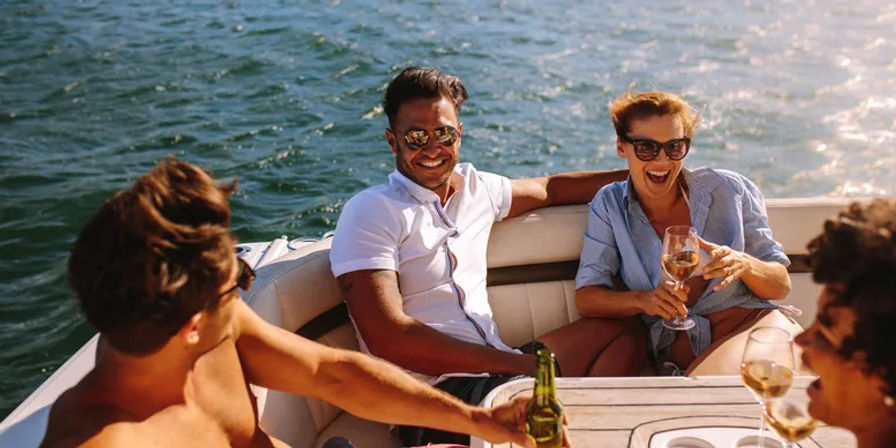 Four friends in sunglasses laughing and toasting with wine and beer on a sunlit motorboat cruising on open water, summer boat party vibe.