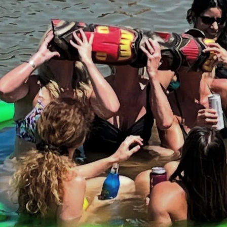 Group of women in swimsuits floating in a sunny lake, hoisting a colorful inflatable tiki log overhead and sipping canned and bottled drinks at a summer water party.
