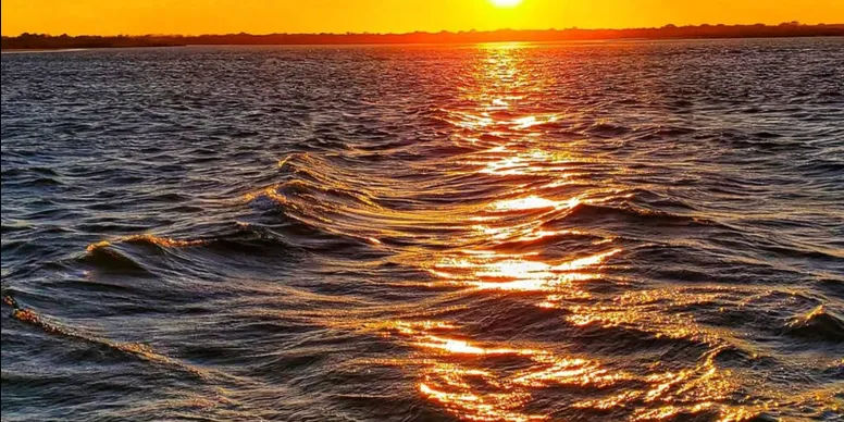 Vibrant orange sunset over open water, the sun casting a golden path across rippling waves toward a low distant shoreline.