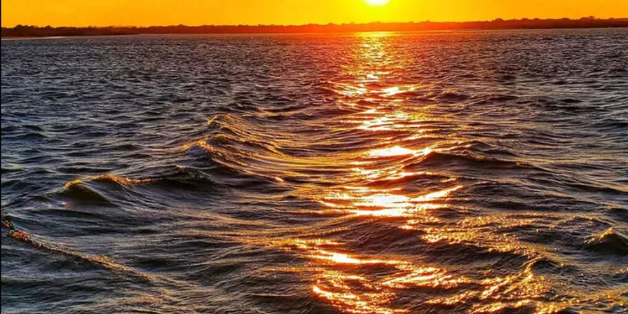 Vibrant orange sunset over open water, the sun casting a golden path across rippling waves toward a low distant shoreline.