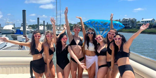 Eight friends in black and white swimsuits and pink heart-shaped sunglasses cheering and posing on a sunny boat at a coastal marina with yachts and waterfront homes in the background.