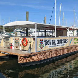 Tiki-themed charter pontoon boat docked at a sunny Florida marina, with wood-grain hull graphics, colorful tiki masks, an orange life ring, and sailboat masts in the background.