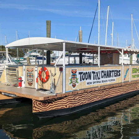 Tiki-themed charter pontoon boat docked at a sunny Florida marina, with wood-grain hull graphics, colorful tiki masks, an orange life ring, and sailboat masts in the background.