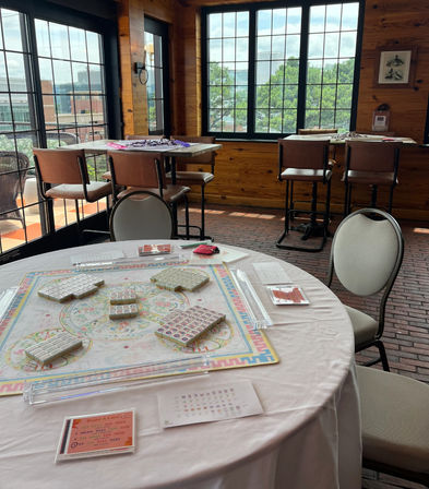 Round table set for mahjong with tiles, racks and score cards on a white tablecloth in a sunny rustic game room with large paned windows, wood-paneled walls, brick floor and high stools overlooking trees and city buildings.