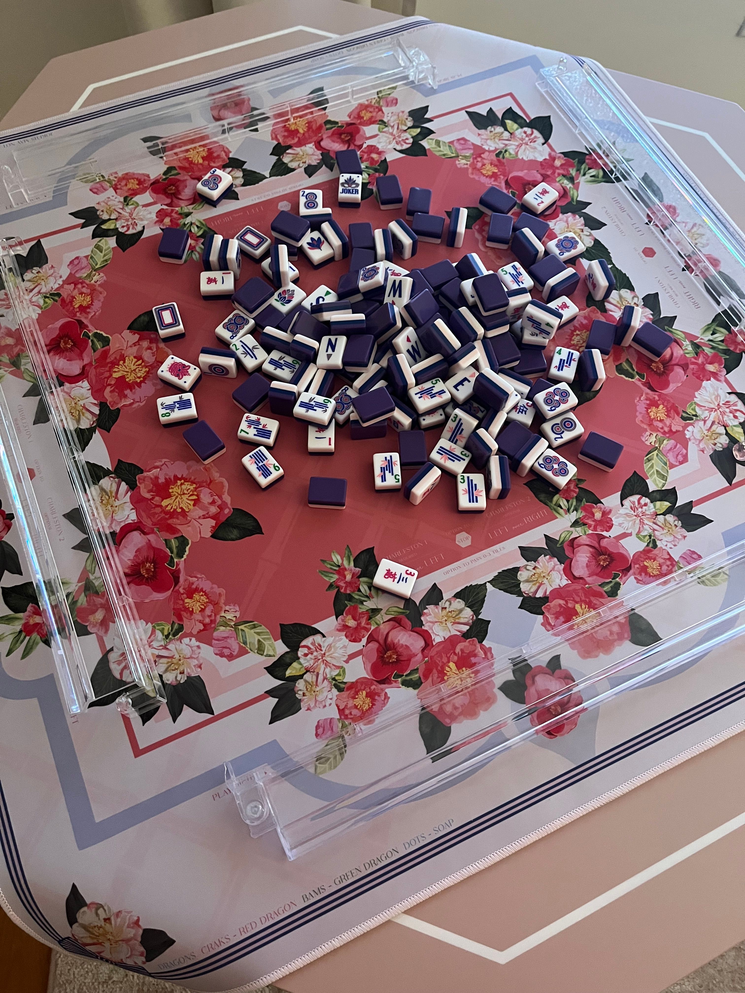 Scattered purple-backed mahjong tiles in a playful pile on a pink floral game mat with clear acrylic racks on a table