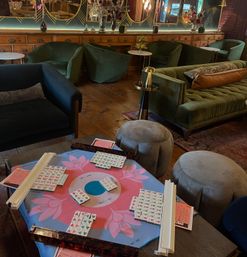 Mahjong set on a pink floral play mat with tiles and racks, surrounded by green velvet sofas, round gray ottomans, wooden floors and decorative mirrors in a cozy lounge