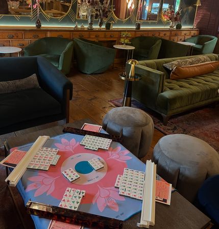 Mahjong set on a pink floral play mat with tiles and racks, surrounded by green velvet sofas, round gray ottomans, wooden floors and decorative mirrors in a cozy lounge