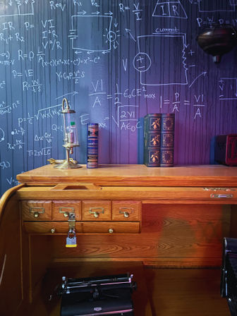 Vintage oak roll-top desk with a locked cubby, antique typewriter, brass lamp and stacked decorative books set against a purple-lit chalkboard wall covered in handwritten physics and circuit equations — quirky science-themed study nook.