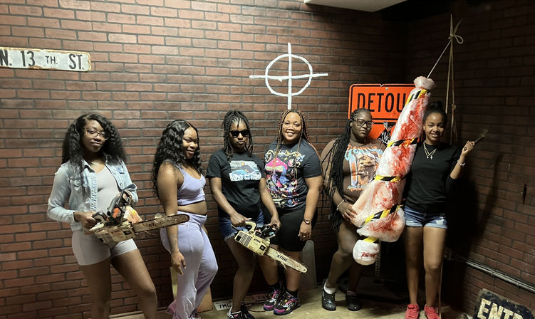 Six women pose in a brick-walled, horror-themed room holding prop chainsaws and a bloodstained wrapped dummy, with a “DETOUR” sign and a “13th St” street sign visible — fun escape-room style group photo.