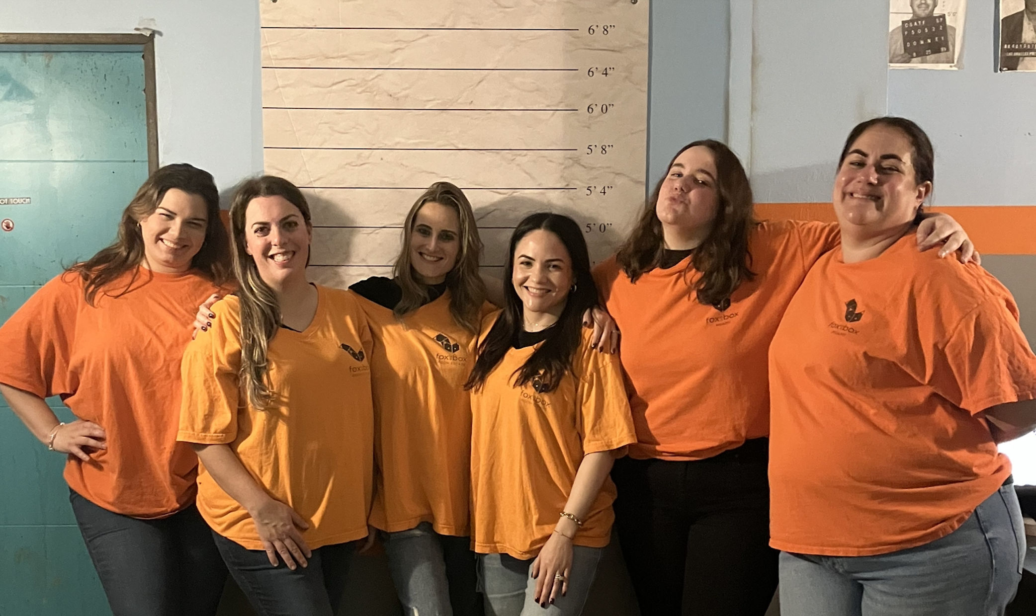 Six women in matching orange shirts and jeans smiling and posing arm-in-arm in front of a mugshot-style height chart on an indoor wall — playful group/team photo.