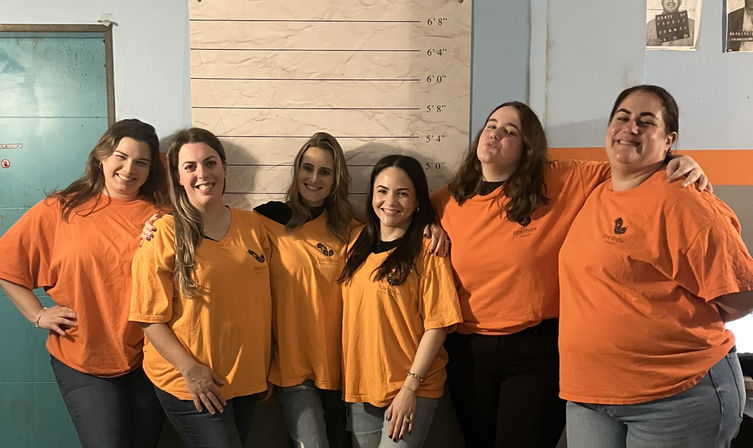Six women in matching orange shirts and jeans smiling and posing arm-in-arm in front of a mugshot-style height chart on an indoor wall — playful group/team photo.