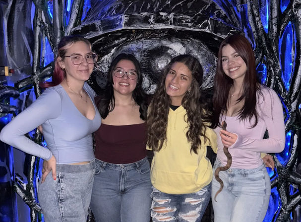Four friends in casual jeans and tops pose and smile in front of a blue-lit sculptural wall at an indoor neon art installation, one holding a small brown snake.