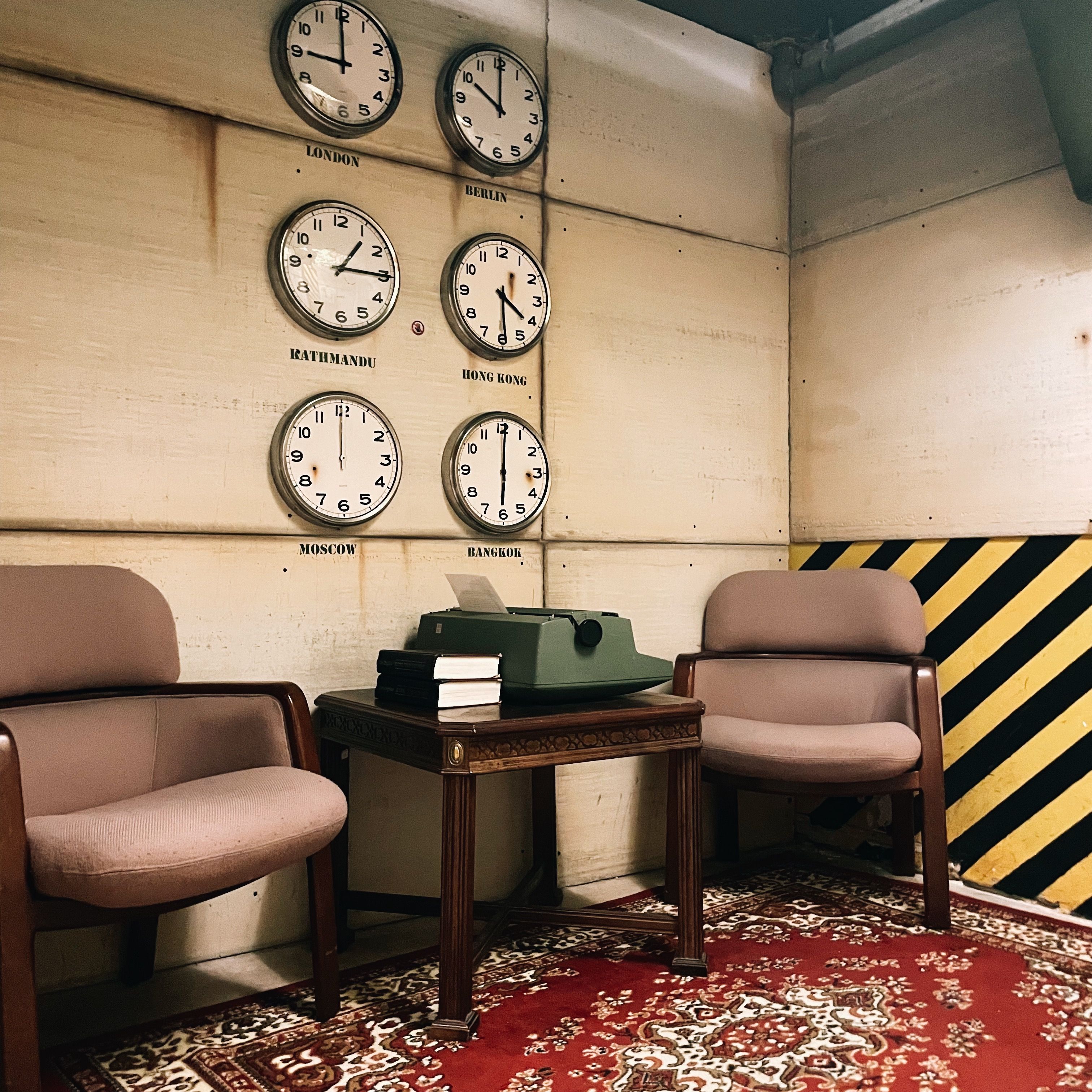 Vintage office corner with six wall clocks labeled London, Berlin, Kathmandu, Hong Kong, Moscow and Bangkok, two mid-century chairs, a wooden side table with a green typewriter and stacked books on a red patterned rug