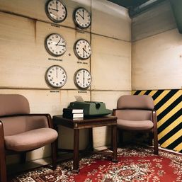 Vintage office corner with six wall clocks labeled London, Berlin, Kathmandu, Hong Kong, Moscow and Bangkok, two mid-century chairs, a wooden side table with a green typewriter and stacked books on a red patterned rug