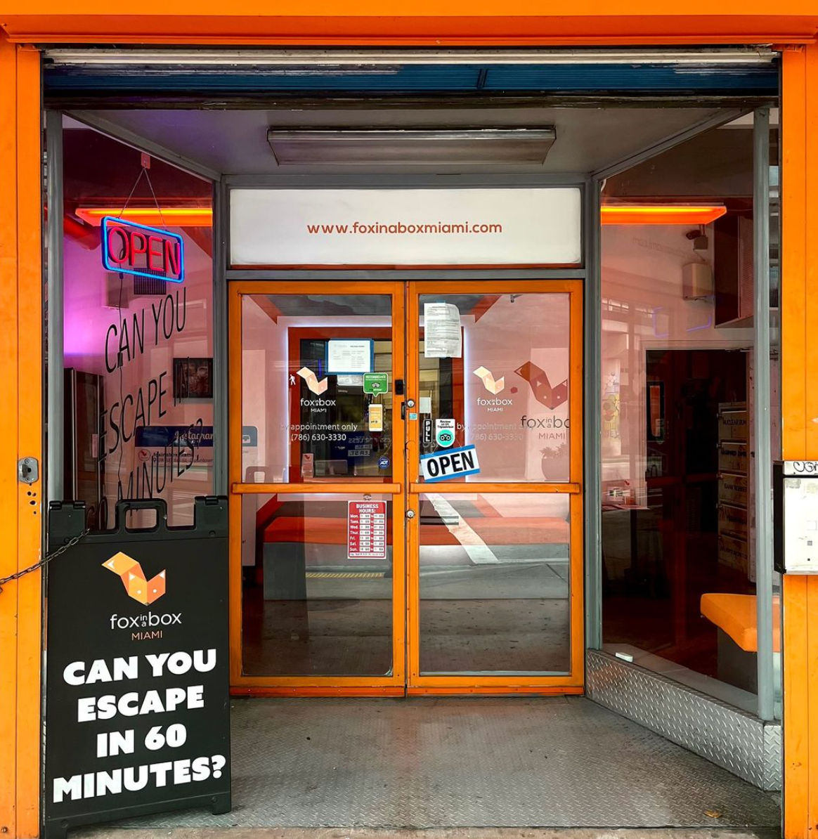 Bright orange escape-room storefront in Miami with glass double doors, neon OPEN sign, and a sidewalk sign asking “Can you escape in 60 minutes?”