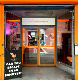 Bright orange escape-room storefront in Miami with glass double doors, neon OPEN sign, and a sidewalk sign asking “Can you escape in 60 minutes?”