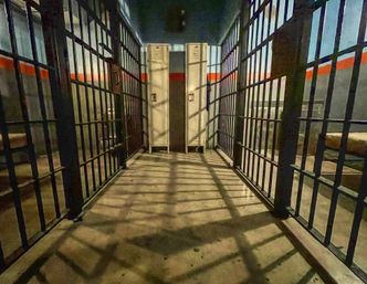 Gritty jail cell corridor interior with steel bars on both sides, twin metal lockers at the end, bunk beds visible and dramatic grid shadows on the concrete floor.