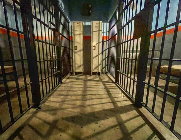 Gritty jail cell corridor interior with steel bars on both sides, twin metal lockers at the end, bunk beds visible and dramatic grid shadows on the concrete floor.