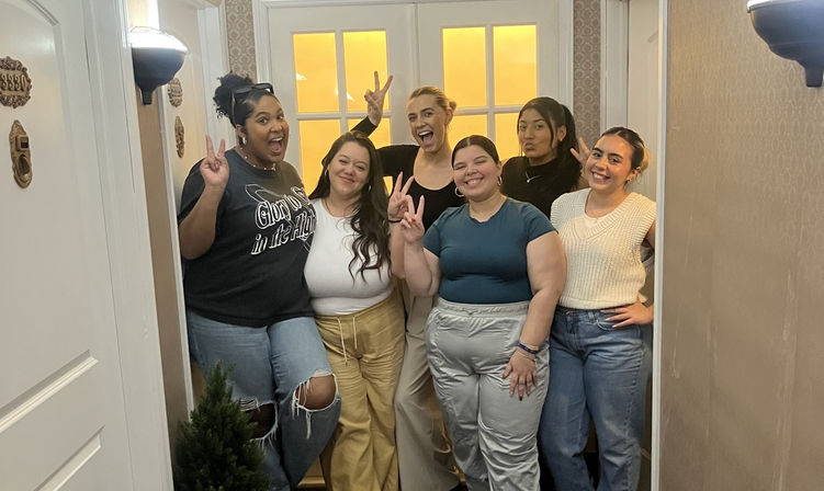 Six friends posing in a narrow indoor hallway by a glass-paneled door, smiling and flashing peace signs in casual outfits and warm lighting.