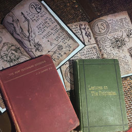 Antique occult display: open illustrated grimoire with arcane symbols and creature sketches beside red and green hardcover books on a woven table