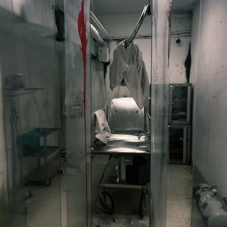 Dim, eerie medical exam room or small morgue with stainless gurney on wheels, hanging white lab coat, blood-red streaks on clear plastic curtains, metal cabinets and scattered equipment