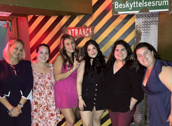 Six women smiling and dressed up, posing together at a nightlife venue in front of a yellow-and-black striped wall and a red "Entrance" sign — friends’ night out group photo.
