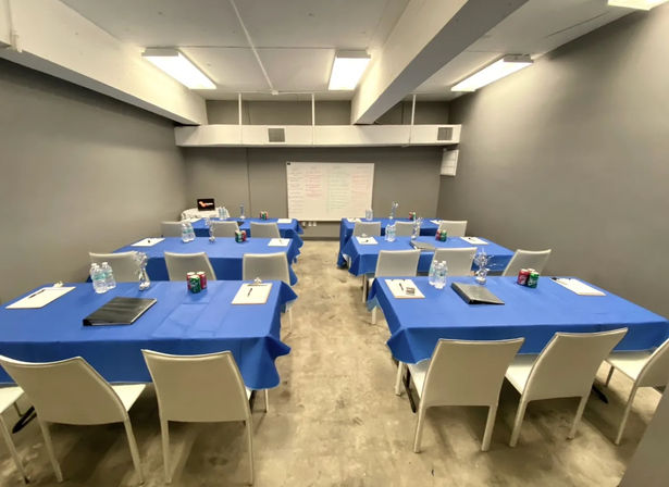 Small indoor training room set up for a workshop — six rectangular tables with blue tablecloths, white chairs, bottled water, notepads and soda cans at each seat, a whiteboard at the front, gray walls and fluorescent ceiling lights.