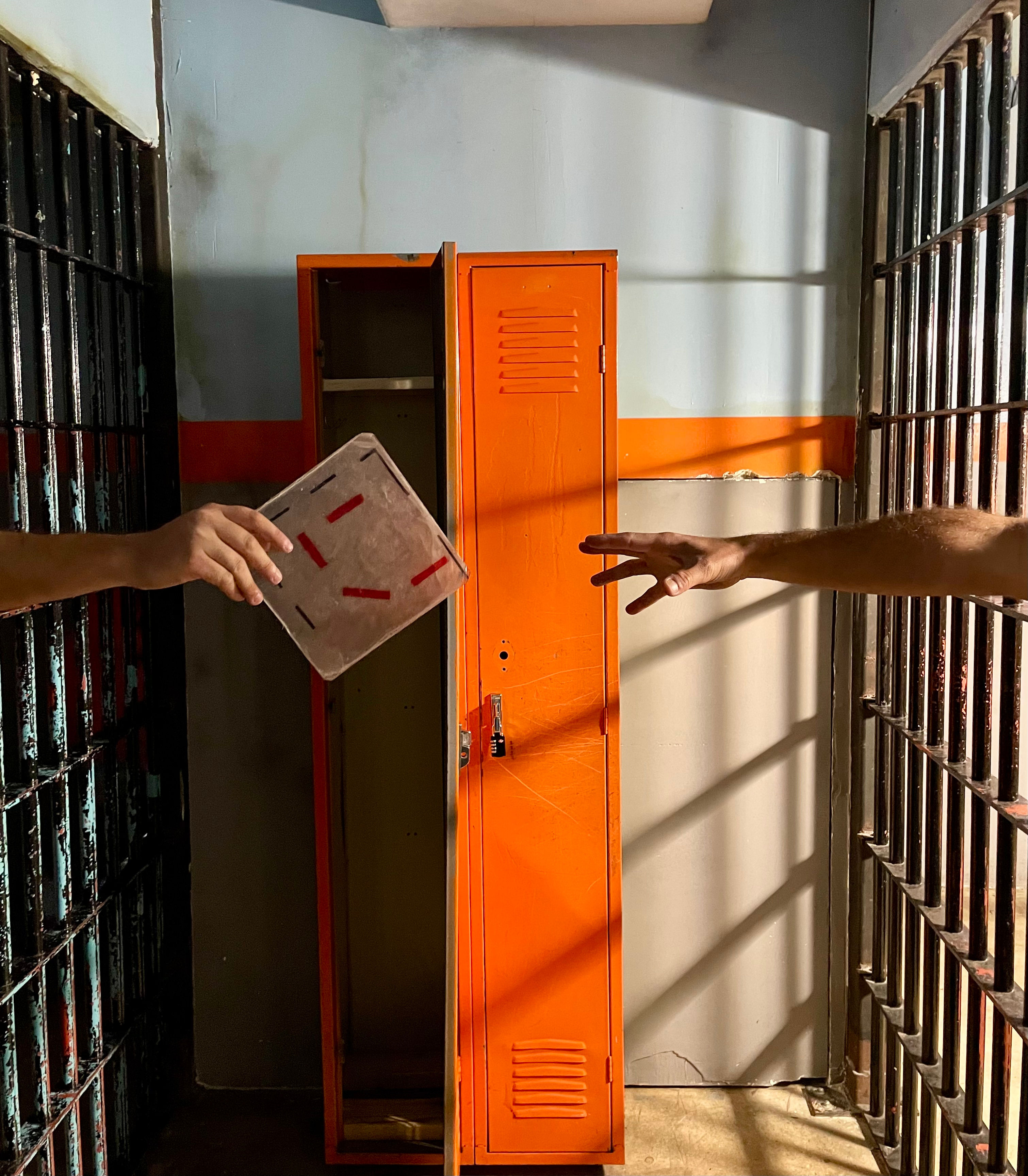 Sunlit jail cell with a bright orange locker between rusted metal bars; two hands reach to pass a small notebook through the gap as striped bar shadows fall on the wall.