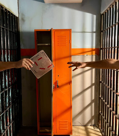 Sunlit jail cell with a bright orange locker between rusted metal bars; two hands reach to pass a small notebook through the gap as striped bar shadows fall on the wall.