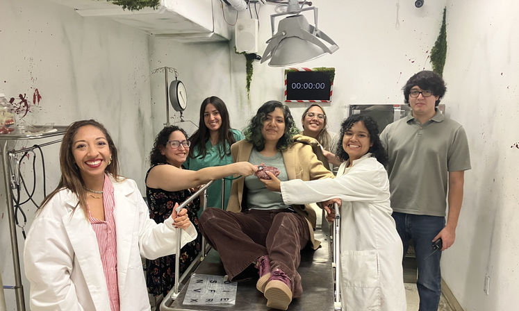 Seven people smiling in a medical-themed escape room: two in lab coats and others gathered around a person sitting on an exam table holding a fake heart prop under surgical lights, with a 00:00:00 digital timer on the wall and blood-spatter decor.