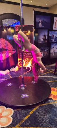 Pole fitness performer in pink outfit doing an inverted hold on a portable pole at an indoor convention booth with purple-pink lighting and photo backdrops.