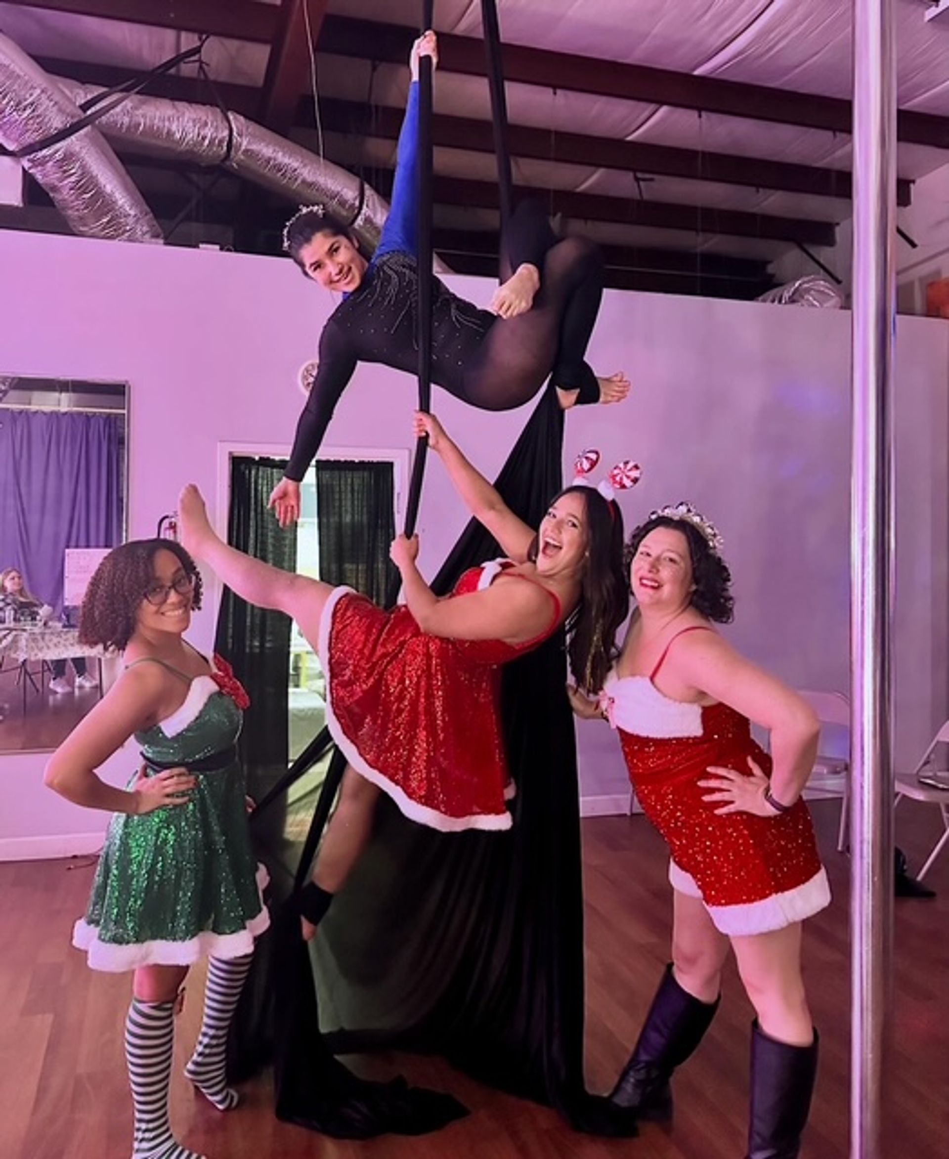 Festive aerial studio shot of four women in holiday costumes — one suspended on aerial silks and a pole, three posing in glittery Santa and elf dresses.
