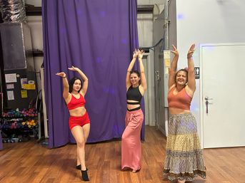 Three women in colorful outfits smiling and striking arms-up dance poses in a hardwood-floor dance studio with a purple curtain backdrop and nearby workout weights — group dance class practice.