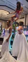 Bridal expo in a hotel ballroom: two women modeling ornate white wedding gowns beside a playful pole performer in pink shorts and crop top under a chandelier