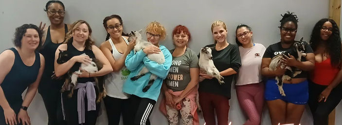 Smiling women posing in an indoor goat yoga class, each holding a playful baby goat in a yoga studio