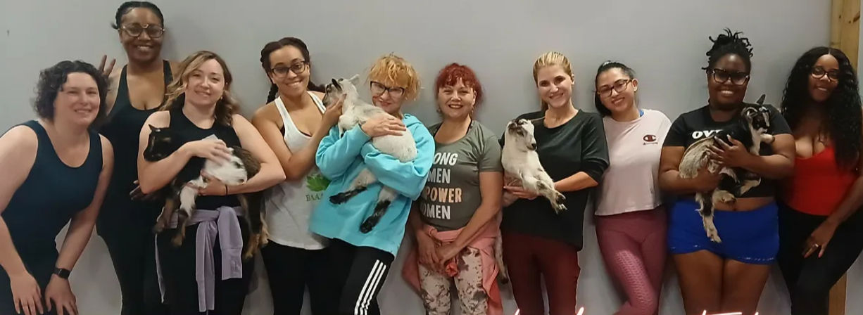 Smiling women posing in an indoor goat yoga class, each holding a playful baby goat in a yoga studio