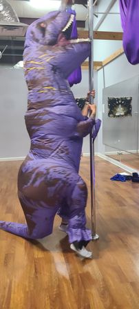 Person in an inflatable purple T‑rex costume playfully holding a pole in a mirrored indoor dance studio with wood flooring