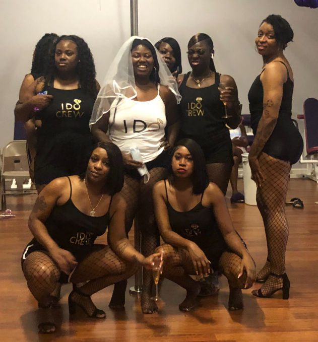 Bachelorette party group photo in a dance studio — bride in a white veil and 'I DO' tank surrounded by friends in matching black 'I DO Crew' outfits and fishnet stockings holding champagne.