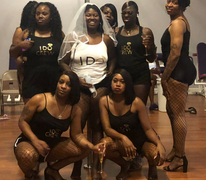 Bachelorette party group photo in a dance studio — bride in a white veil and 'I DO' tank surrounded by friends in matching black 'I DO Crew' outfits and fishnet stockings holding champagne.