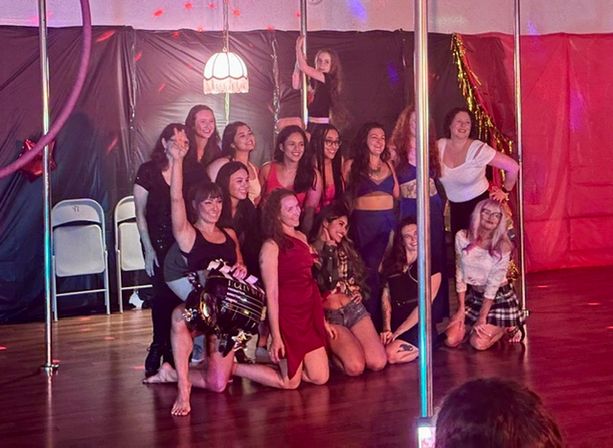 Group of women posing for a celebratory photo in an indoor pole-fitness studio with chrome poles, warm stage lighting and a vintage hanging lamp, some in activewear and dresses on a wooden floor.